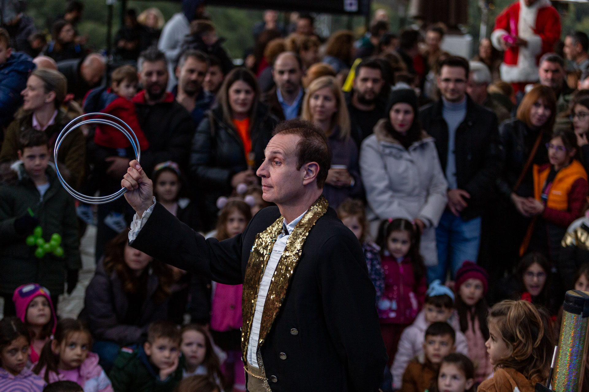 Ένας μάγος που φοράει ένα χρυσό σακάκι κρατάει στα χέρια του πολλούς κρίκους και πλήθος παιδιών και γονέων γύρω του τον παρακολουθούν
