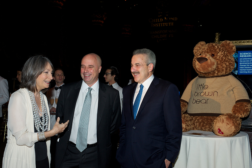 Ο Αντρέας Δρακόπουλος, Πρόεδρος του Ιδρύματος Σταύρος Νιάρχος, μιλάει με μια γυναίκα και έναν άντρα και χαμογελούν και δίπλα τους υπάρχει ένα τραπέζι στο οποίο είναι τοποθετημένος ένας λούτρινος αρκούδος