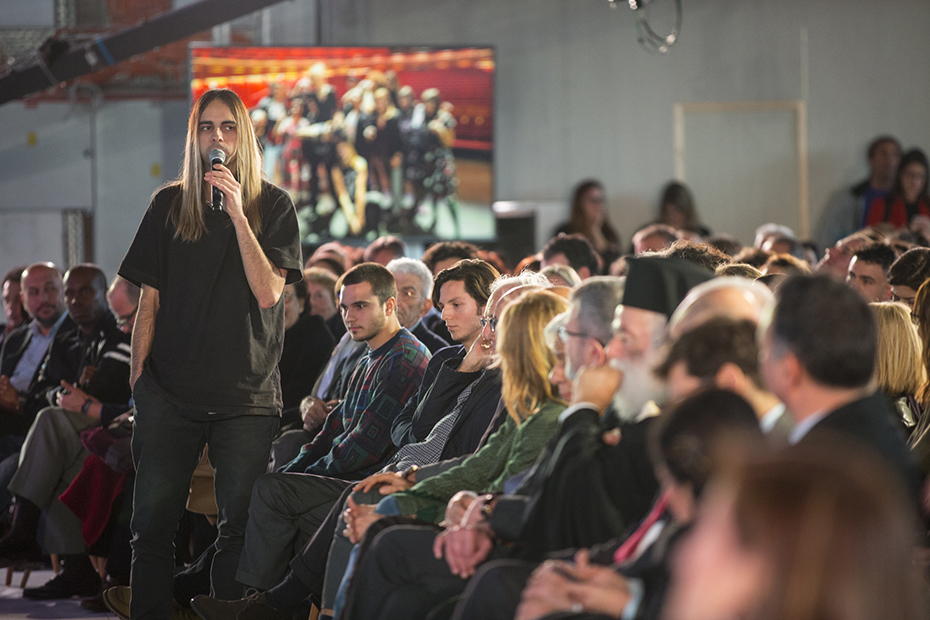 Ένας νεαρός από το κοινό σηκώνεται και θέτει έναν προβληματισμό στο μικρόφωνο καθώς το υπόλοιπο κοινό τον κοιτά 