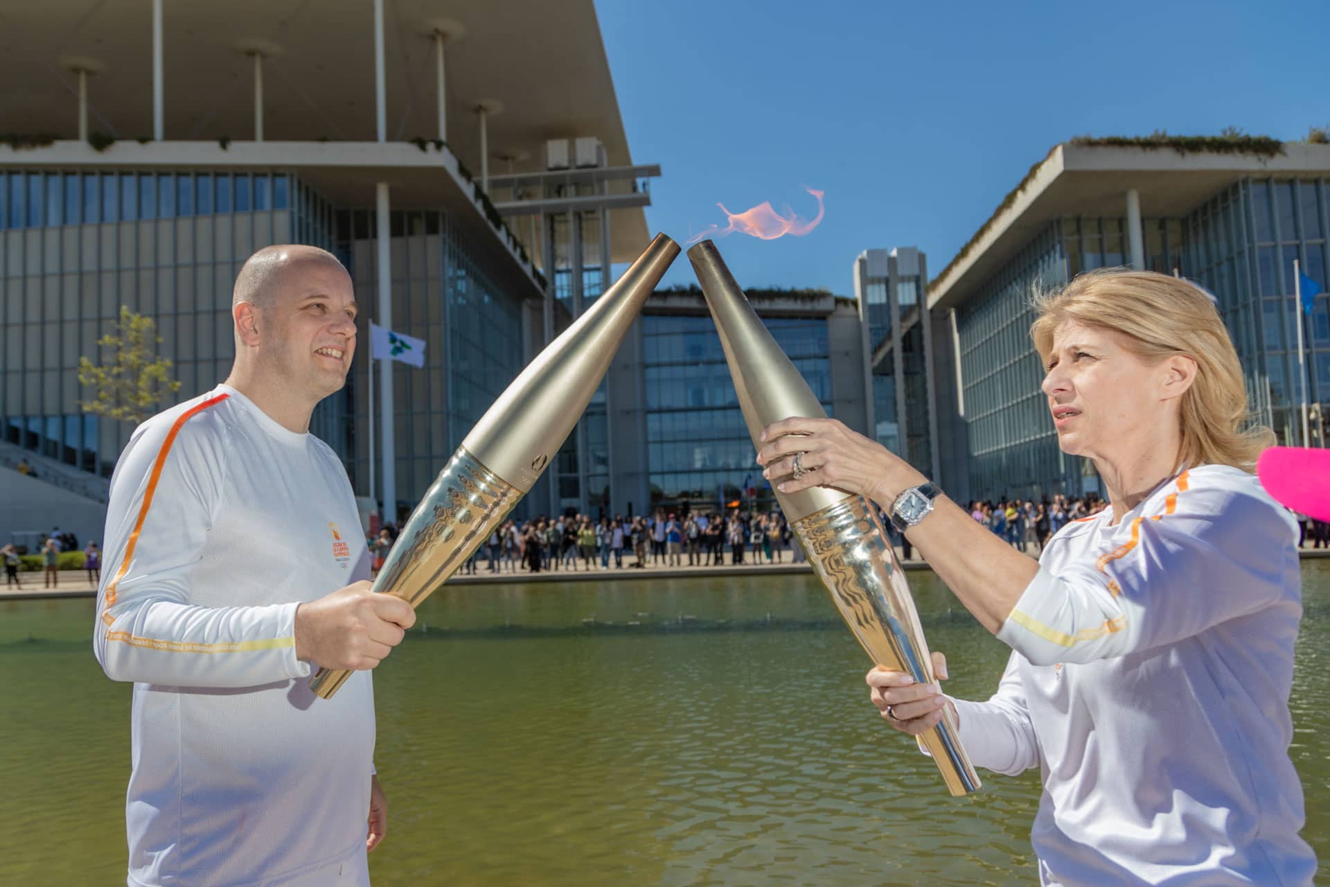 Ένας άνδρας και μια γυναίκα, που κρατούν ο καθένας μια μοντέρνα δάδα και περνούν τη φλόγα από τον έναν στον άλλον μπροστά από το κανάλι του Κέντρου Πολιτισμού Ίδρυμα Σταύρος Νιάρχος.