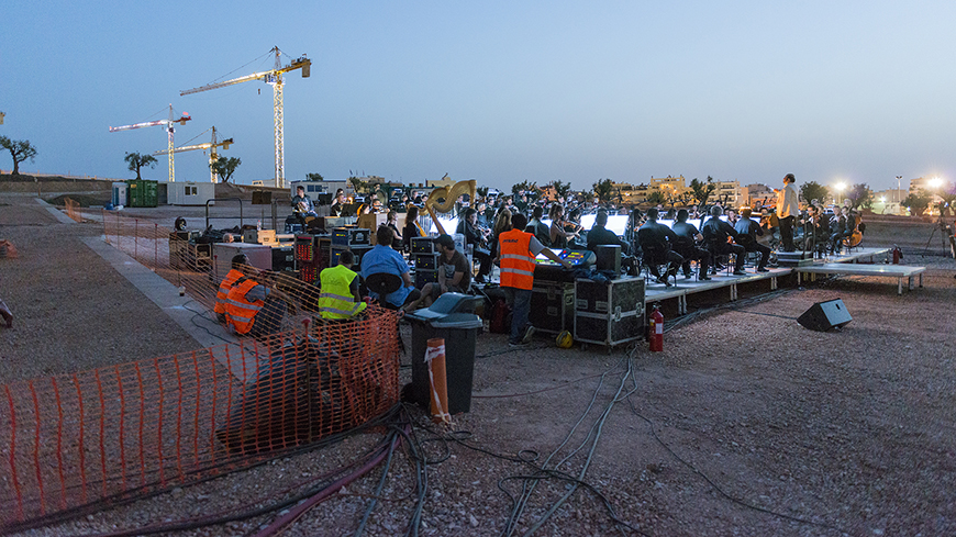 Μια σκηνή τοποθετημένη στο κέντρο ενός εργοταξίου πάνω στην οποία κάθονται μέλη μιας ορχήστρας με τα όργανά τους