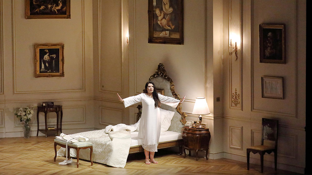 Μια γυναίκα στέκεται όρθια με τα χέρια ανοιχτά φορώντας ένα λευκό νυχτικό μπροστά από ένα κρεβάτι το οποίο βρίσκεται μέσα σε ένα μεγάλο και εμβληματικό δωμάτιο 