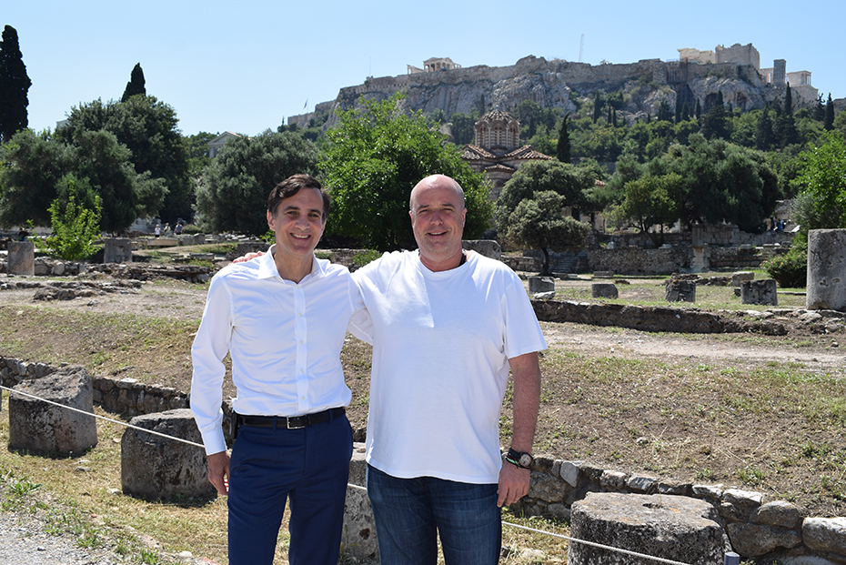 Ο Ανδρέας Δρακόπουλος και ένας άλλος άντρας χαμογελούν και κουνιούνται και βγάζουν μια φωτογραφία έξω από έναν αρχαιολογικό χώρο με την Ακρόπολη στο βάθος