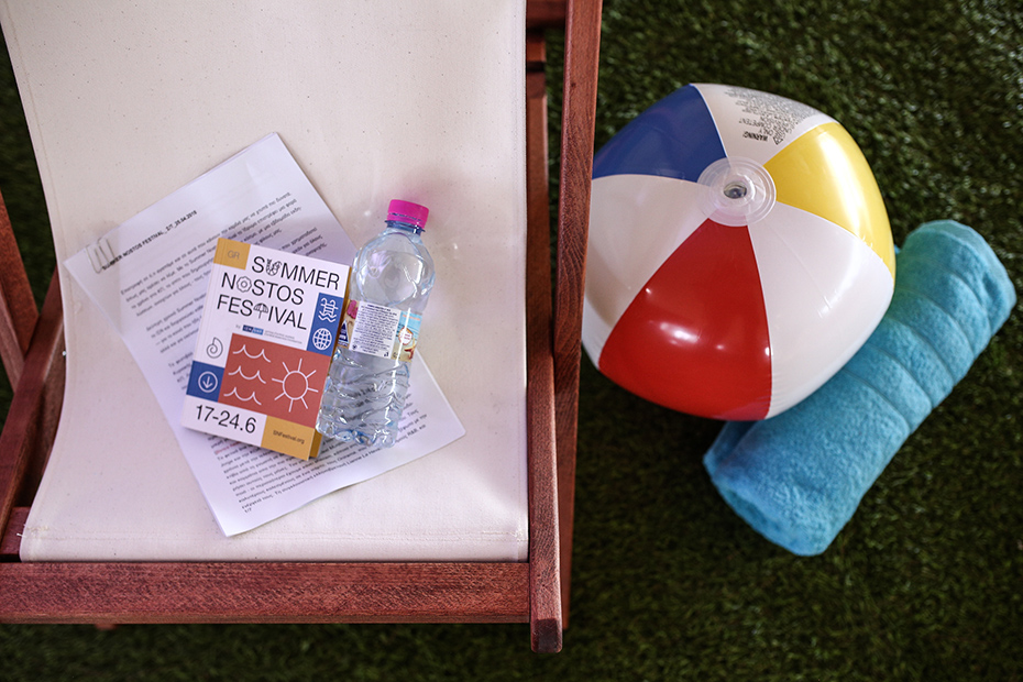 Ένα φύλλο χαρτί, ένα φυλλάδιο του Summer Nostos Festival και ένα μπουκάλι νερό πάνω σε μια καρέκλα γκαζόν, δίπλα σε μια μπάλα θαλάσσης και μια πετσέτα.