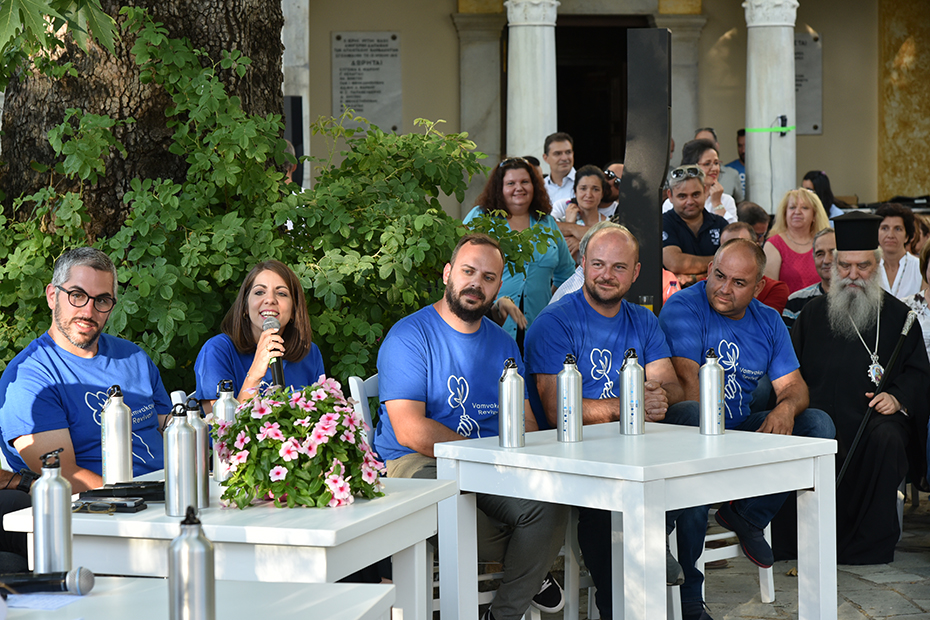 Φωτογραφία από μια ομάδα ανθρώπων που φοράει το ίδιο μπλουζάκι, να κάθεται στην κεντρική πλατεία του χωριού και μια γυναίκα από την ομάδα να κρατάει το μικρόφωνο και να μιλάει