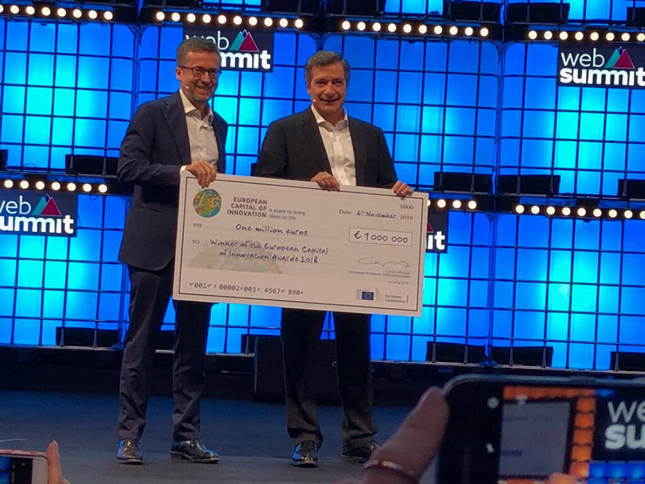 Δύο άντρες πάνω στη σκηνή κρατάνε στα χέρια τους μια επιταγή σε μέγεθος μακέτας και χαμογελούν 