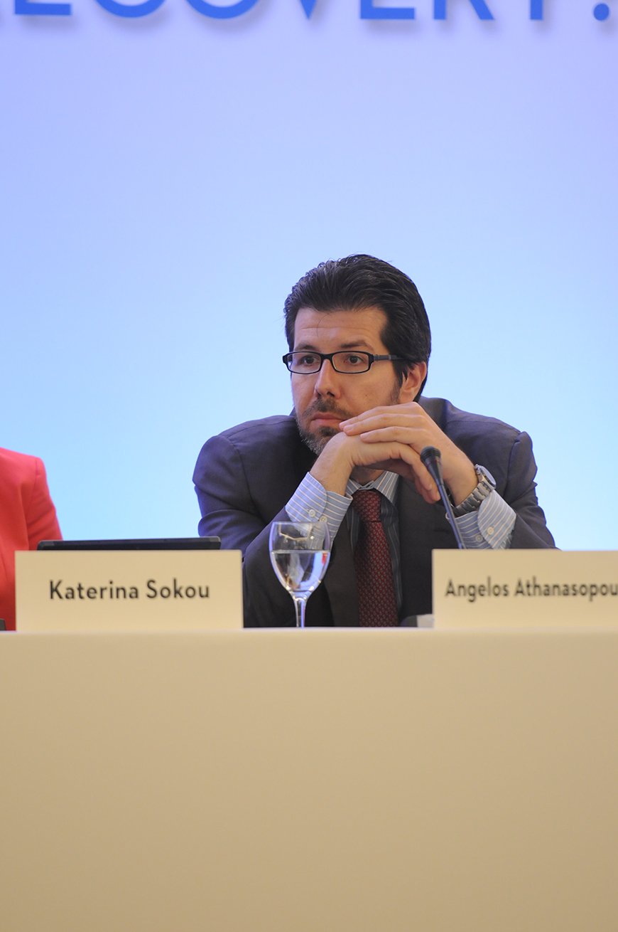 Κοντινή φωτογραφία ενός άντρα ο οποίος κάθεται σε ένα τραπέζι και έχει μπροστά του ένα μικρόφωνο και ένα καρτελάκι που γράφει το όνομά του, Angelos Athanasopoulos 