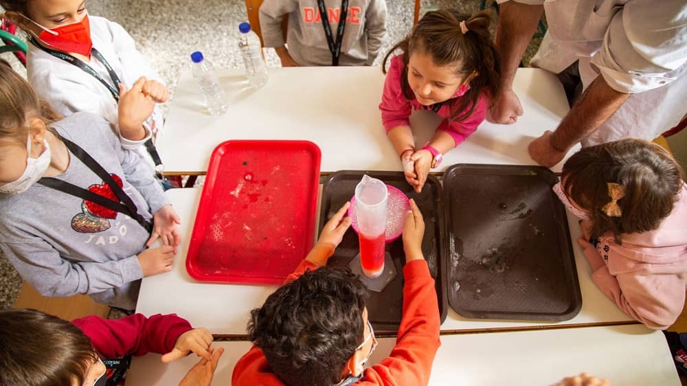 Πολλά παιδιά συγκεντρώνονται γύρω από ένα τραπέζι και κάνουν ένα πείραμα