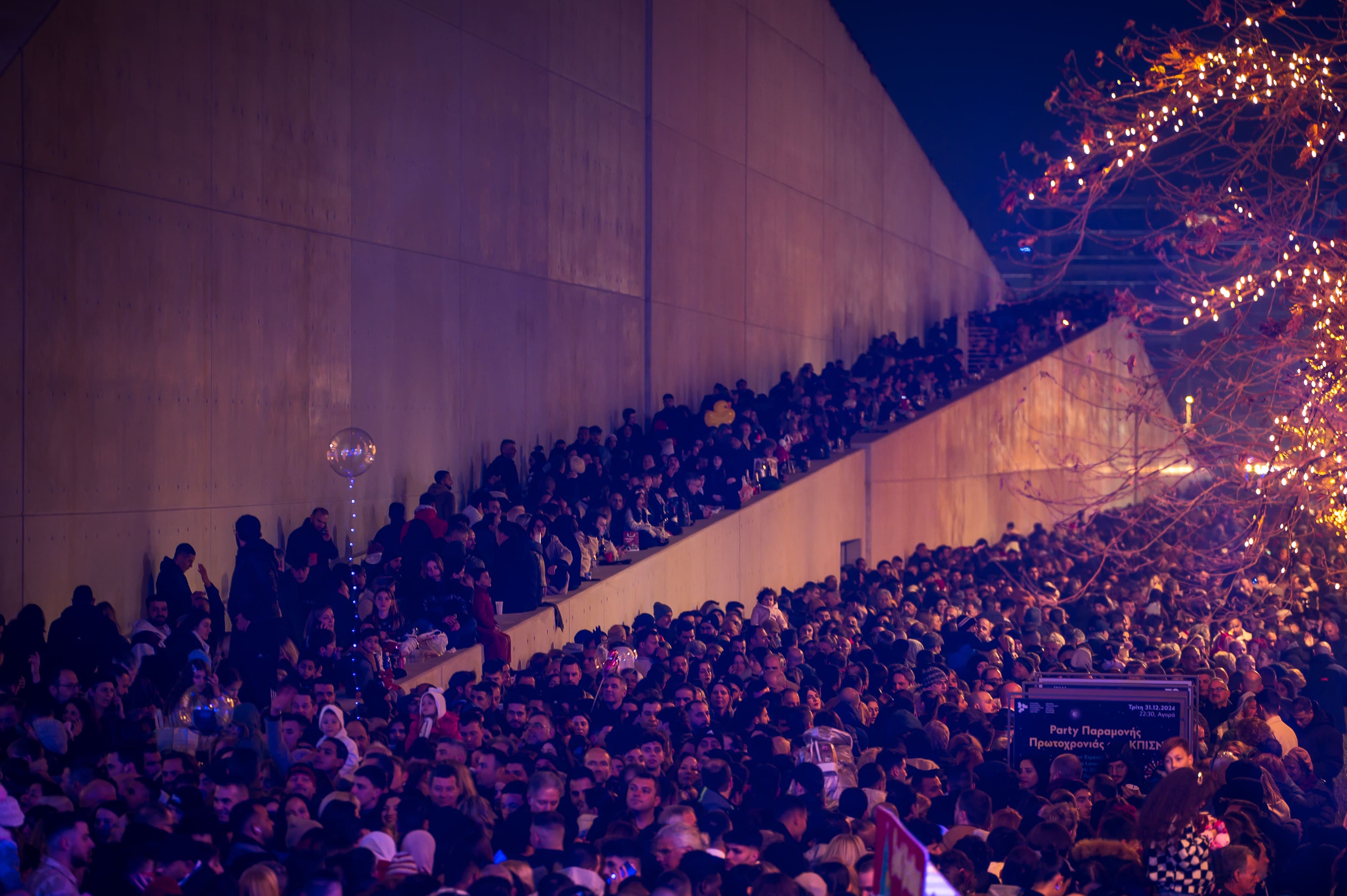 Μεγάλο πλήθος που στέκεται στη σκάλα της Αγοράς του ΚΠΙΣΝ, δίπλα από ένα φωτισμένο δέντρο.