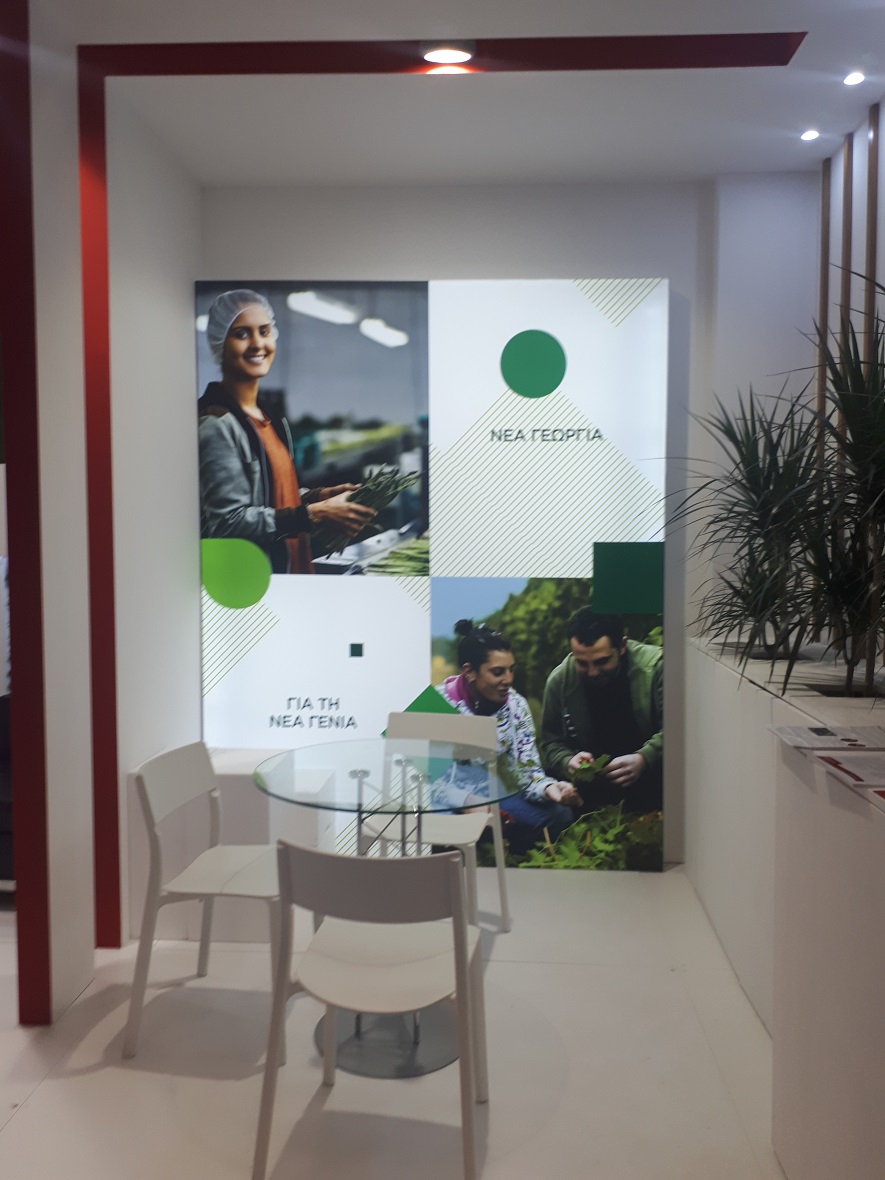 Μια γωνία ενός εσωτερικού χώρου στον οποίο υπάρχει ένα μικρό στρογγυλο τραπέζι με τρεις καρέκλες και στον τοίχο υπάρχει ένα μεγάλο banner που γράφει ΝΕΑ ΓΕΩΡΓΙΑ ΓΙΑ ΤΗ ΝΕΑ ΓΕΝΙΑ