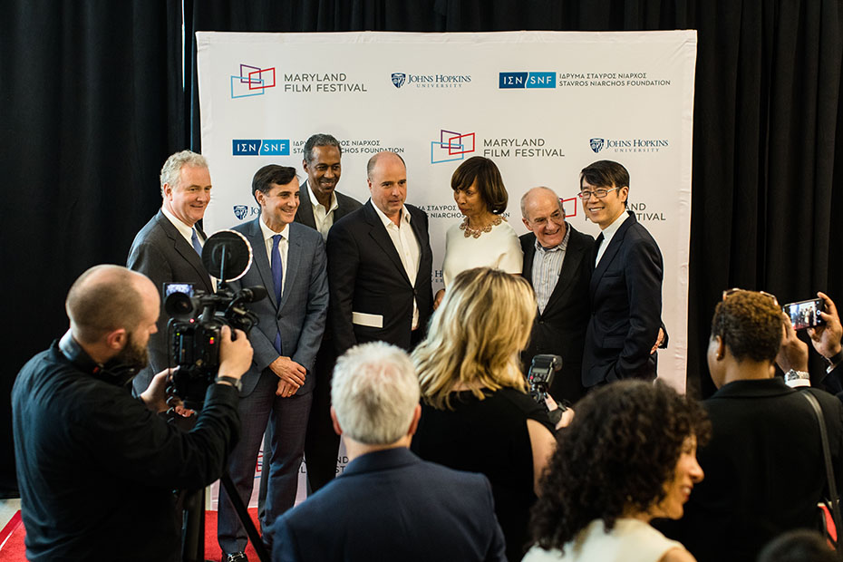 Άνθρωποι φωτογραφίζονται μπροστά από ένα μεγάλο banner το οποίο έχει τα logo του Ιδρύματος Σταύρος Νιάρχος, του John Hopkins University και του MARYLAND FILM FESTIVAL 