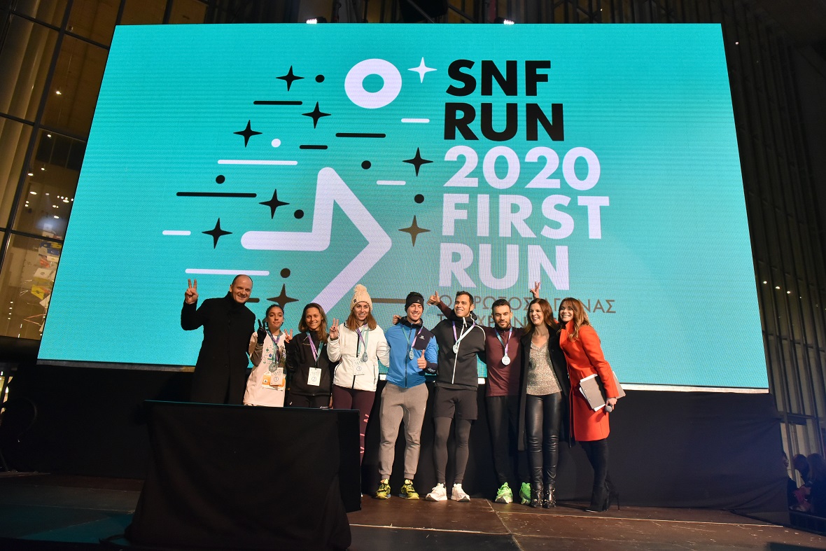 Μια ομάδα ανθρώπων στέκεται μπροστά από μια τεράστια φωτεινή επιγραφή που γράφει SNF RUN 2020 FIRTS RUN για να φωτογραφιθούν 
