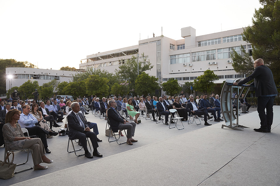 Φωτογραφία που δείχνει πλήθος ανθρώπων να κάθονται σε καρέκλες σε έναν προαύλιο χώρο το οποίο παρακολουθεί τον άντρα που μιλάει στο βήμα 