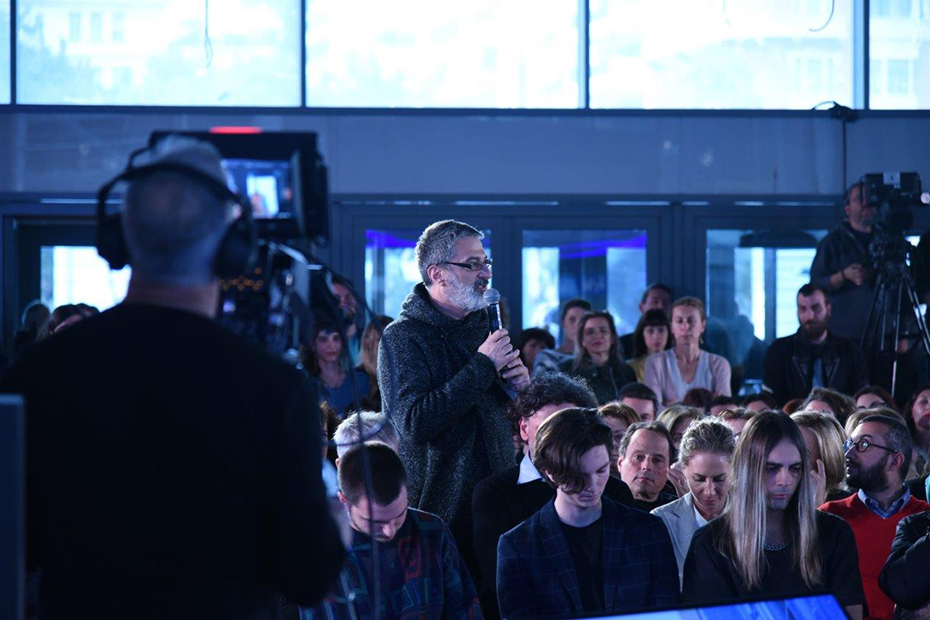 Φωτογραφία που δείχνει έναν βιντεογράφο να βγάζει βίντεο έναν άντρα που στέκεται όρθιος ανάμεσα στο κοινό και μιλάει στο μικρόφωνο 