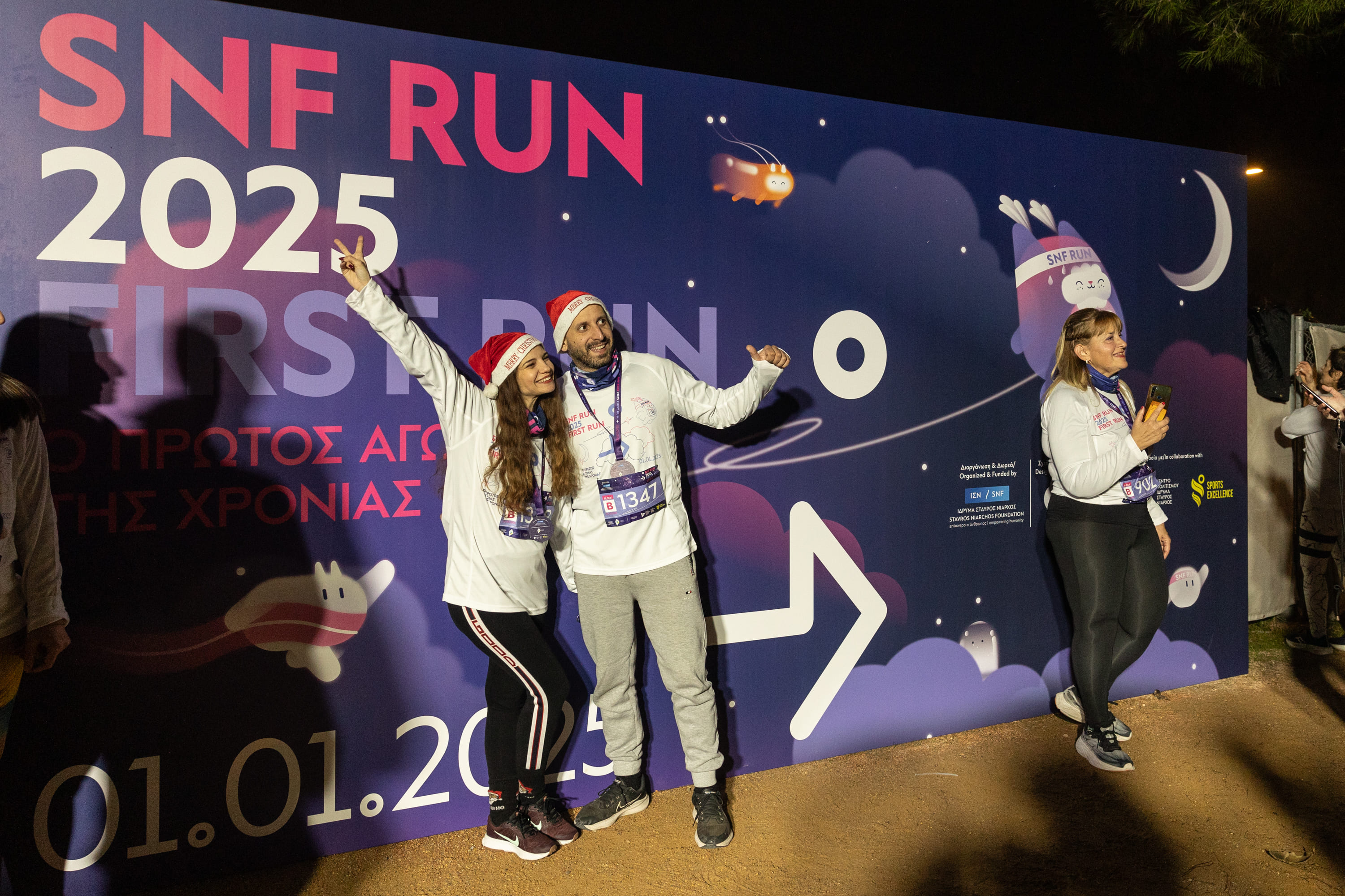 Άτομα που ποζάρουν μπροστά από το πανό του SNF First Run φορώντας χριστουγεννιάτικα καπέλα.