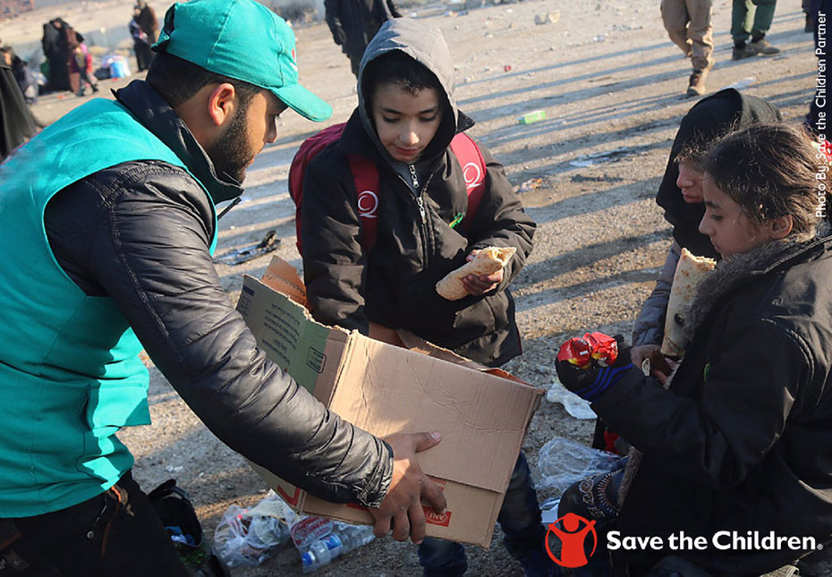 Ένας άντρας κρατάει μια κούτα στα χέρια του και προσφέρει από τα πράγματα τηυς κούτας σε μικρά παιδιά