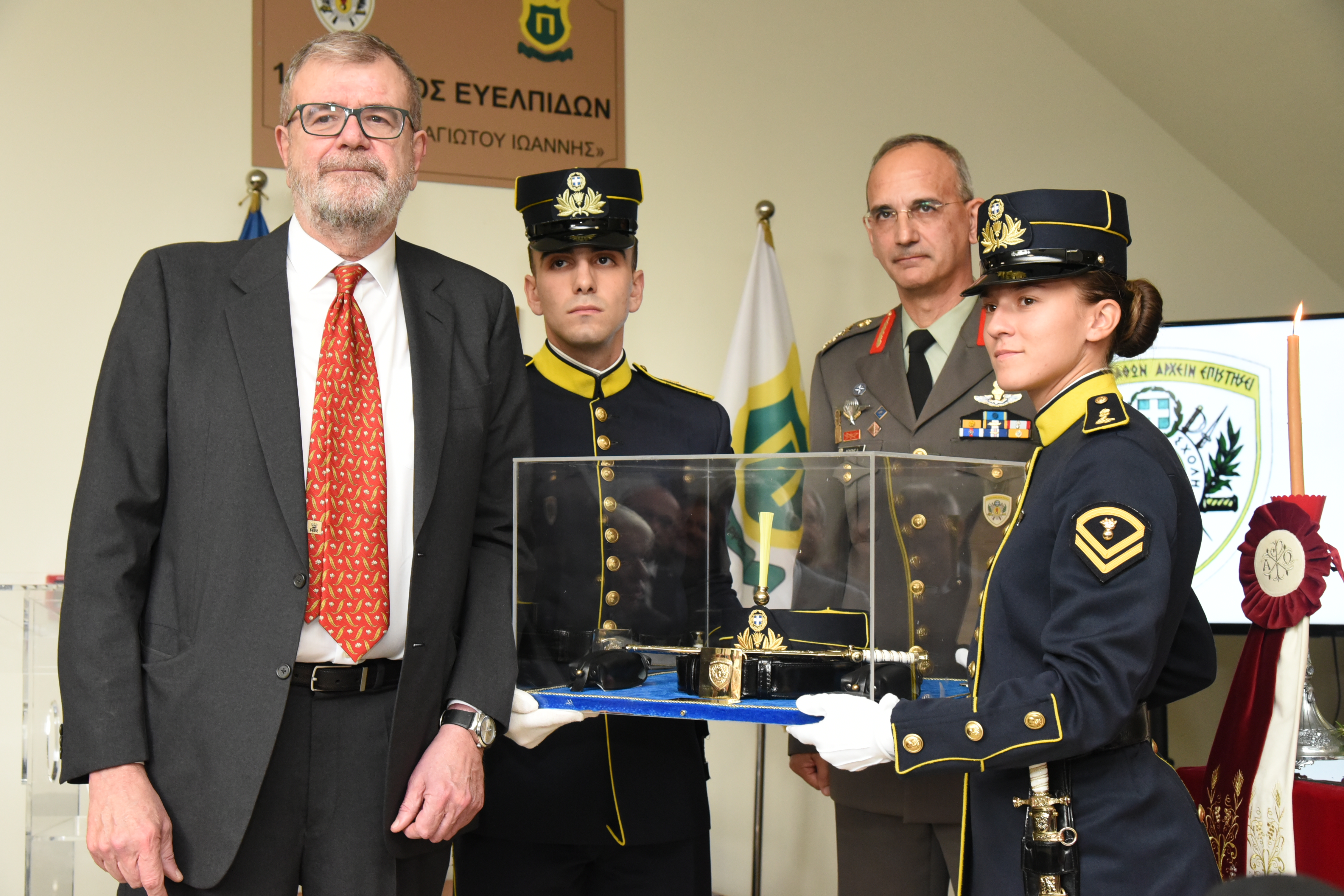 Ένας άνδρας στέκεται μπροστά από έναν νεαρό άνδρα και μια γυναίκα με στρατιωτική ενδυμασία, οι οποίοι κρατούν ένα αντικείμενο κλεισμένο σε γυαλί