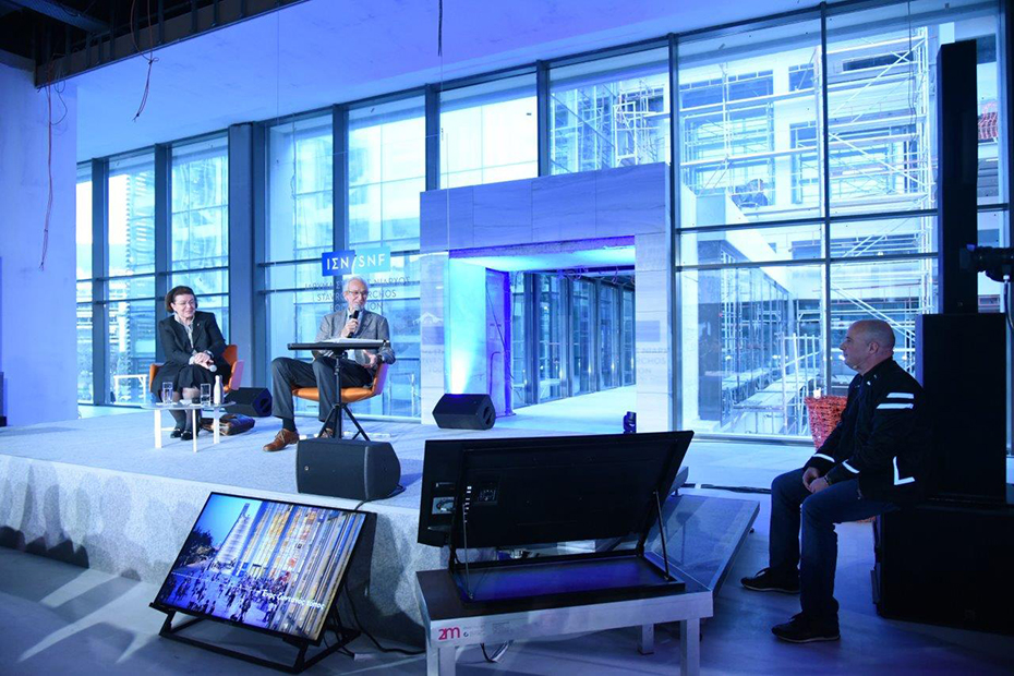 Φωτογραφία που δείχνει έναν άντρα και μια γυναίκα να κάθονται στη σκηνή και ο άντρας μιλάει στο μικρόφωνο και κοιτάει έναν άντρα ο οποίος κάθεται κάτω από τη σκηνή 