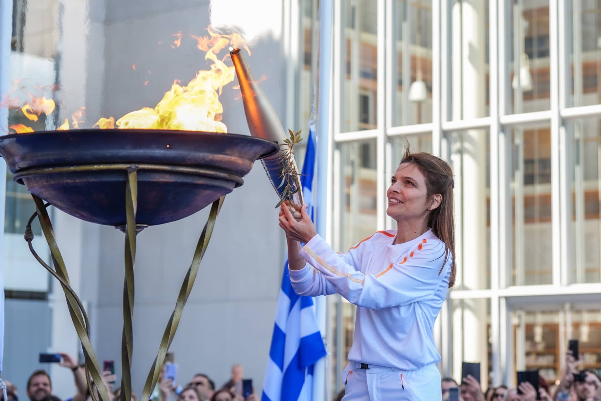 Μια γυναίκα που κρατάει μια μοντέρνα δάδα και ένα κλαδί ελιάς χρησιμοποιεί τη δάδα για να ανάψει μια μεγάλη φλόγα σε έναν τρίποδα, καθώς το πλήθος βιντεοσκοπεί με κινητά τηλέφωνα.