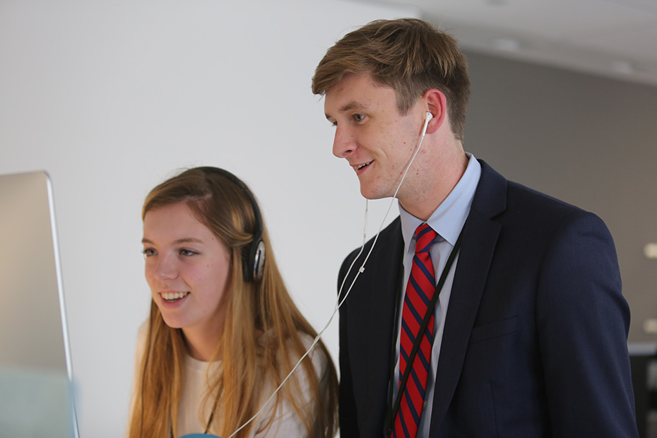 Ένας νεαρός άντρας και μια νεαρή γυναίκα στέκονται δίπλα μπροστά από μια οθόνη υπολογιστή και φοράνε ακουστικά 