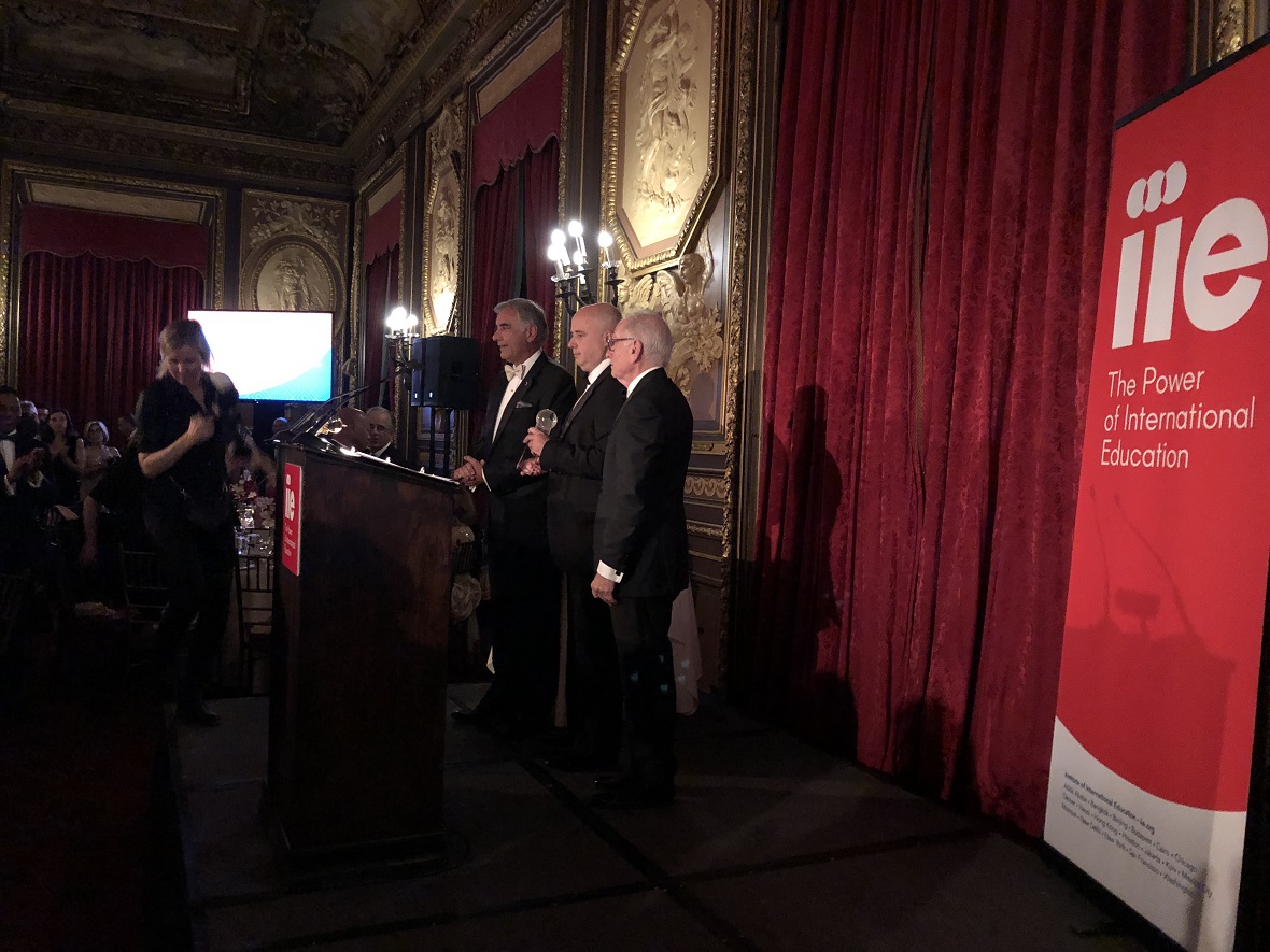 Φωτογραφία τραβηγμένη από το πλαι η οποία δείχνει τον Αντρέα Δρακόπουλο, Πρόεδρο του Ιδρύματος Σταύρος Νιάρχος μαζί με άλλους δυο άντρες στη σκηνή να παραλαμβάνει ένα βραβείο 