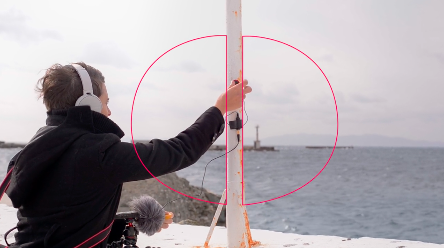 A person holding audio equipment places a microphone onto a pole in front of the sea