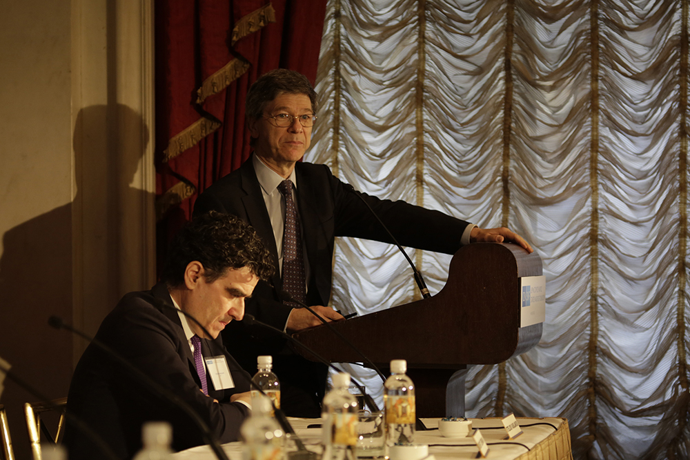 Ένας άντρας κάθεται σε ένα τραπέζι ενώ ένας άντρας δίπλα του μιλάει σε ένα βήμα