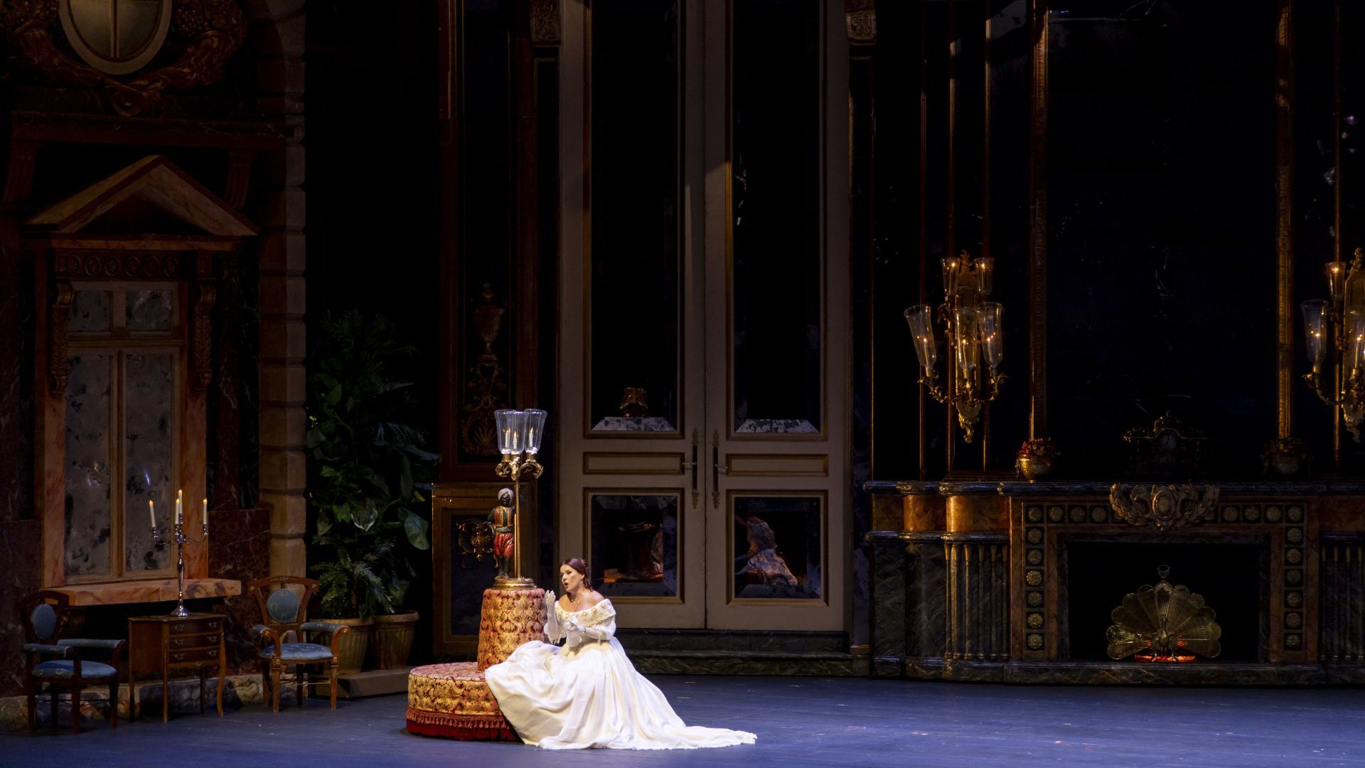 A soprano in an impressive white gown sits on a sofa, in front of a set resembling a palace hall.