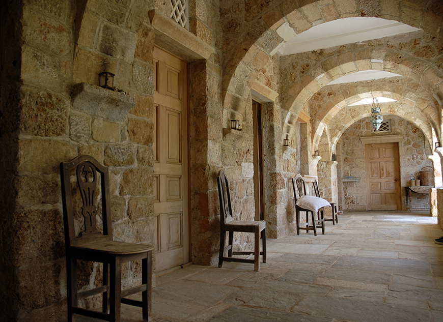 Φωτογραφία που δείχνει έναν διάδρομο ενός πέτρινου οικίματος  με πολλές πόρτες όπου μπροστά από την κάθε μια υπάρχει μια καρέκλα