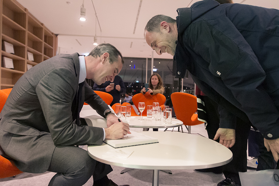 Δύο άντρες σκυμμένοι πάνω από ένα τραπέζι χαμογελάνε όσο ο ένας υπογράφει ένα βιβλίο 