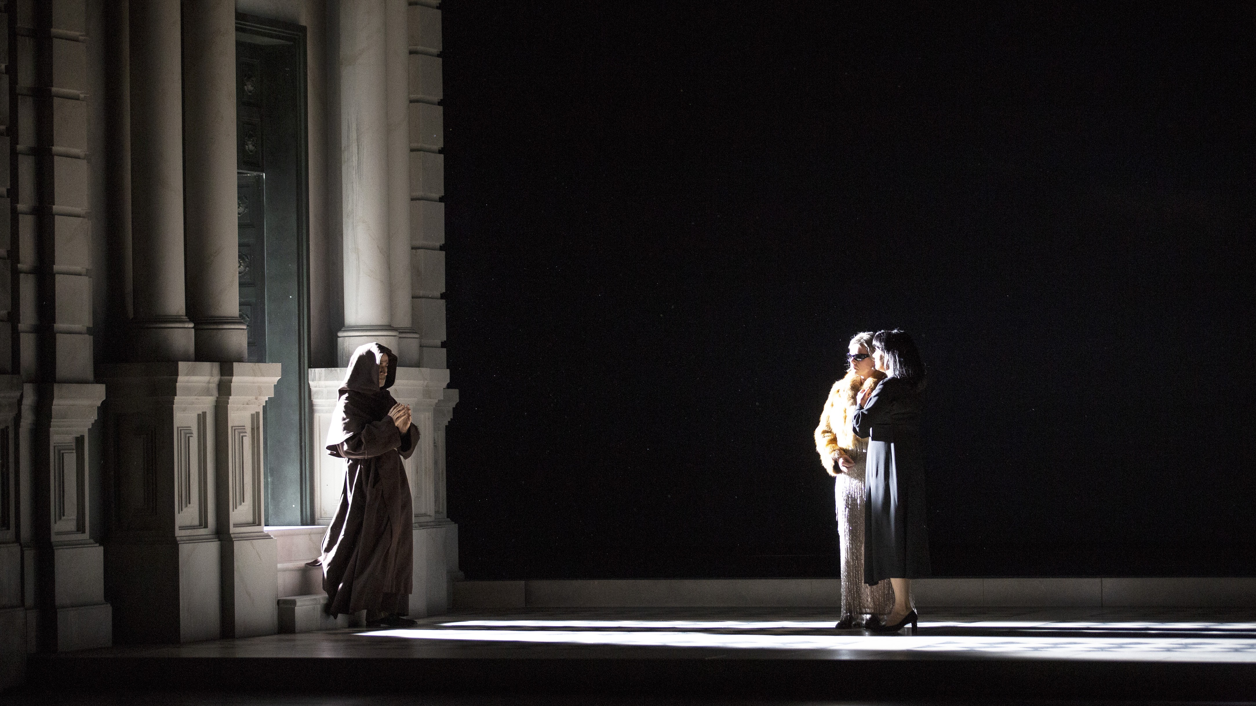 Two women stand by a window, dimly lit in a dark room, while a figure in a black hooded robe stands across them.
