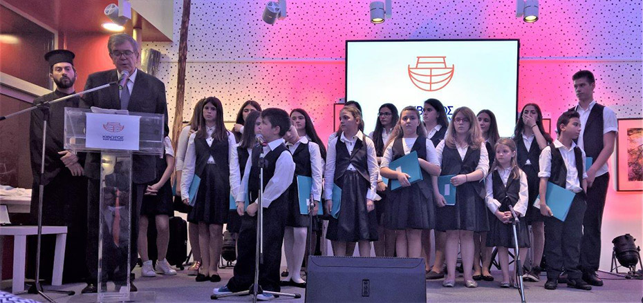 Ένας άντρας μιλάει στο βήμα της ΚΙΒΩΤΟΥ και δίπλα του τα παιδιά της χορωδίας τον παρακολουθούν 