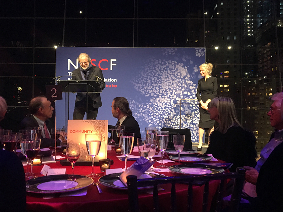 Φωτογραφία που δείχνει το στρογγυλό τραπέζι από το οποίο παρακολουθεί το κοινό τον άντρα που βρίσκεται στο βήμα στη σκηνή και τη γυναίκα που βρίσκεται στην άκρη της σκηνής 