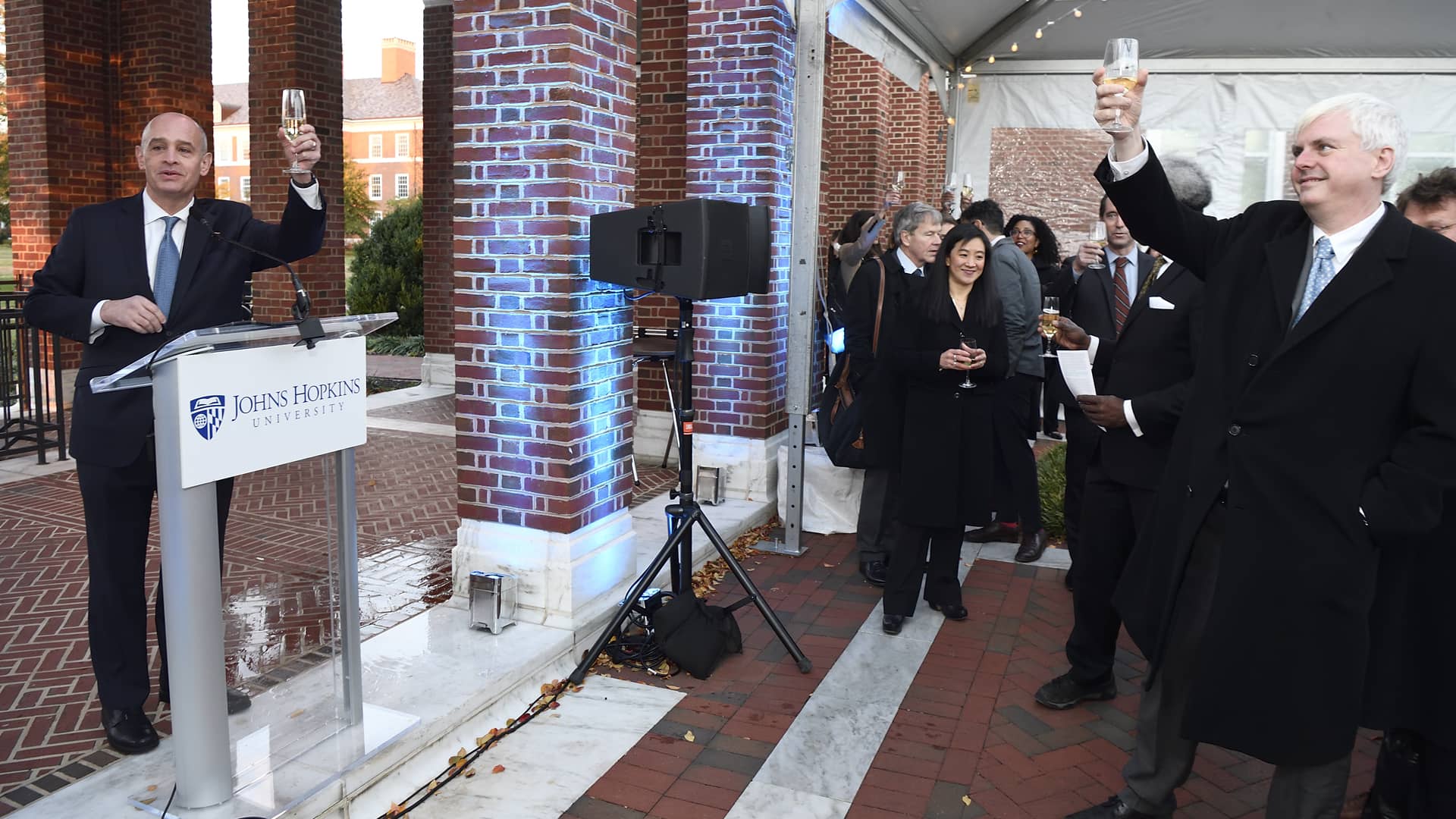 Δύο άντρες που στέκονται απέναντι ο ένας από τον άλλο σηκώνουν το χέρι τους ψιλά κρατώντας ένα ποτήρι σαμπάνια