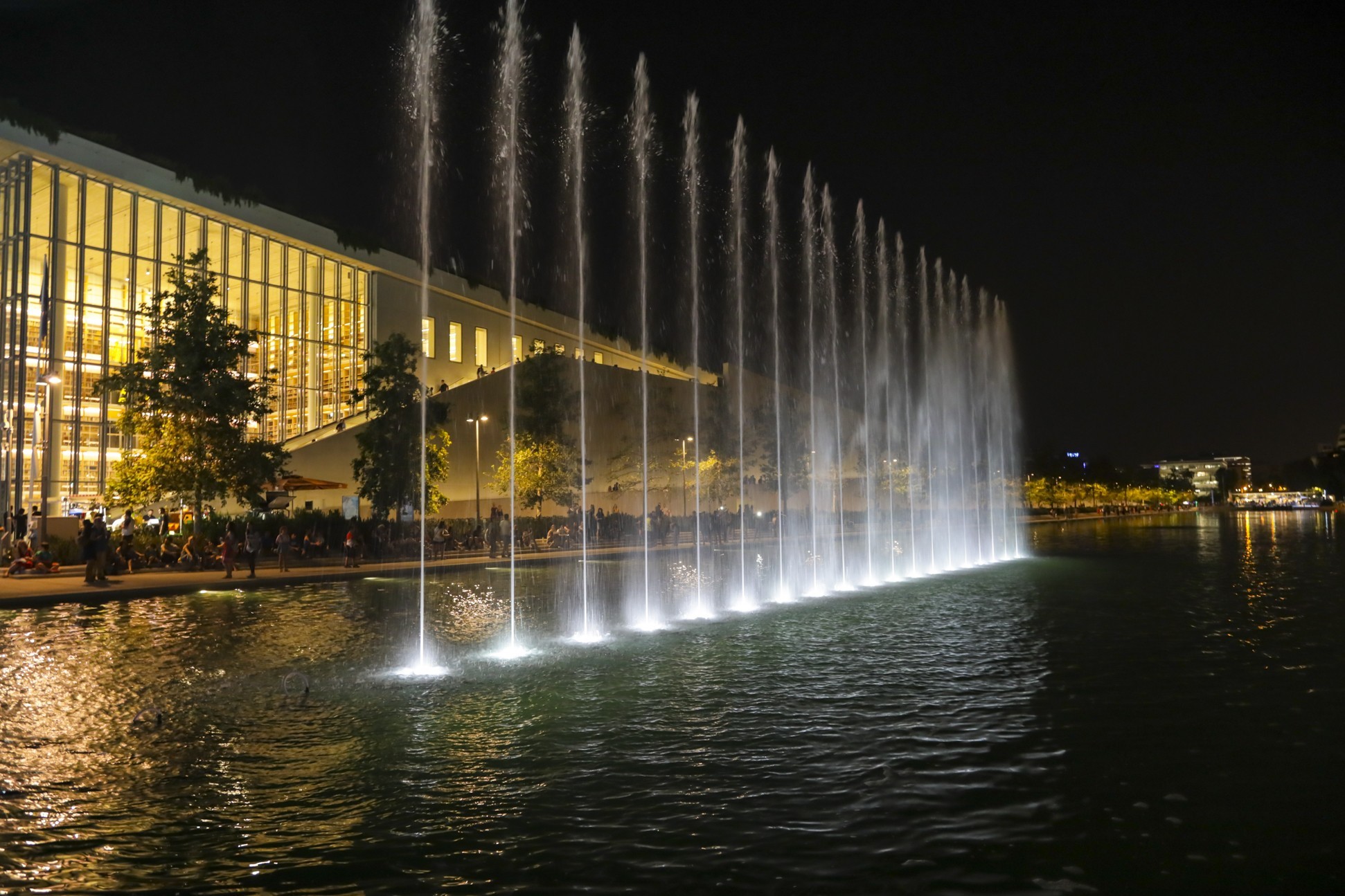 Φωτογραφία από το show που κάνει το συντριβάνι στο κέντρο μιας λίμνης μπροστά από ένα μεγάλο γυάλινο κτήριο 