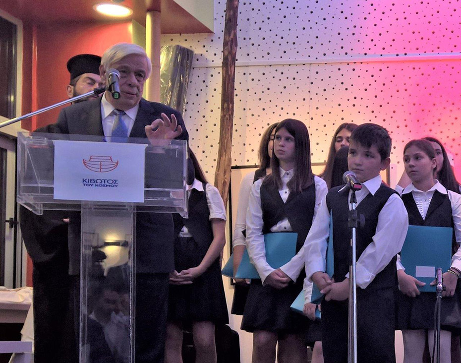Ο Προκόπης Παυλόπουλος, μιλάει στο βήμα της ΚΙΒΩΤΟΥ, ενώ δίπλα του υπάρχει μια ομάδα παιδιών που είναι μέλη της χορωδίας