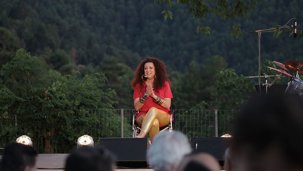 Μια γυναίκα πάνω στη σκηνή κάθεται σε μια καρέκλα και μιλάει στο κοινό 