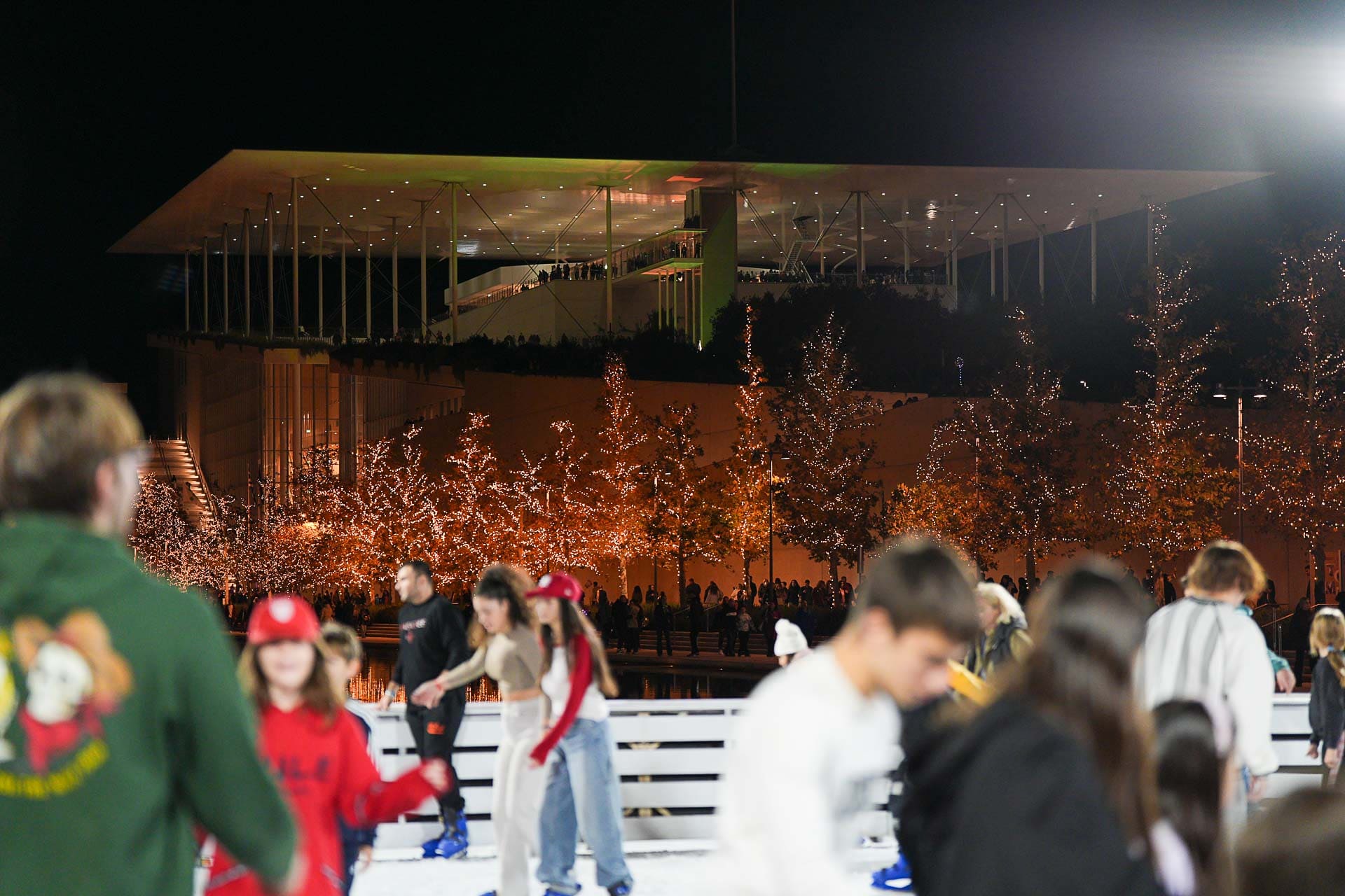 Άτομα που κάνουν πατινάζ στο παγοδρόμιο μπροστά από το κτίριο του ΚΠΙΣΝ και μια σειρά φωτισμένων δέντρων.