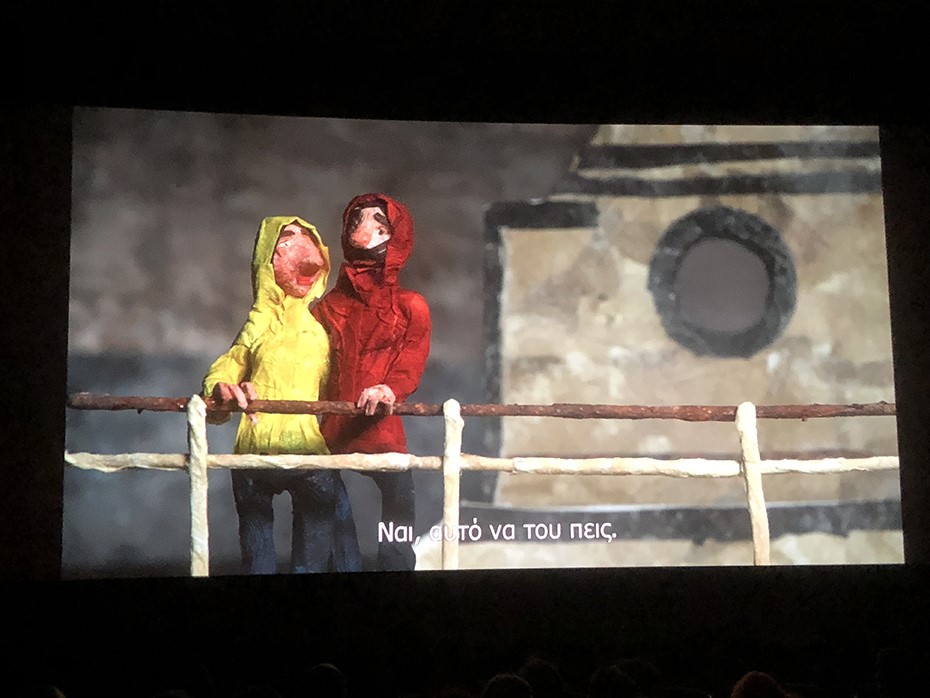 Φωτογραφία στην οποία φαίνεται μια μεγάλη οθόνη του σινεμά στην οποία προβάλλεται με τη μορφή καρτούν δύο φίλοι να μιλούν πάνω σε μια γέφυρα 