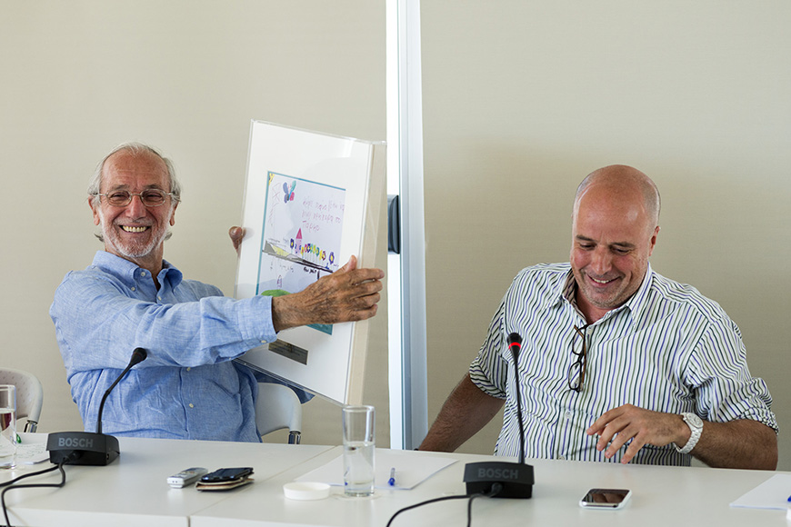 Ο Αντρέας Δρακόπουλος, Πρόεδρος του Ιδρύματος Σταύρος Νιάρχος, κάθεται με έναν άλλον άντρα σε ένα τραπέζι με μικρόφωνα ο οποίος κρατάει ψηλά ένα μεγάλο βιβλίο όπου στο εξώφυλλο υπάρχει μια παιδική ζωγραφιά και χαμογελούν