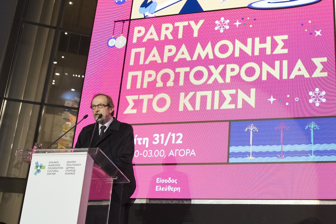 Ένας άντρας στέκεται στο βήμα του Κέντρου Πολιτισμού Ίδρυμα Σταύρος Νιάρχος και πίσω του υπάρχει ένα φόντο που γράφει PARTY ΠΑΡΑΜΟΝΗΣ ΠΡΩΤΟΧΡΟΝΙΑΣ ΣΤΟ ΚΠΙΣΝ