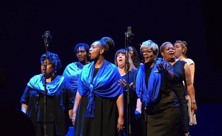 Μια ομάδα γυναικών ομοιμόρφα ντυμένη στέκεται στη σκηνή και τραγουδά 