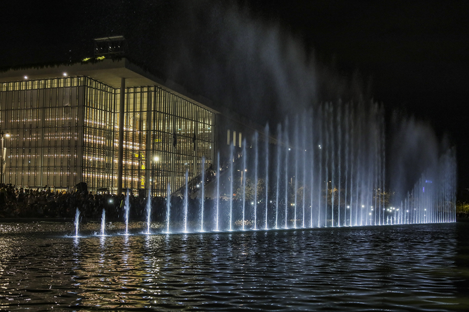 Φωτογραφία από το show που κάνει το συντριβάνι στο κέντρο μιας λίμνης μπροστά από ένα μεγάλο γυάλινο κτήριο 