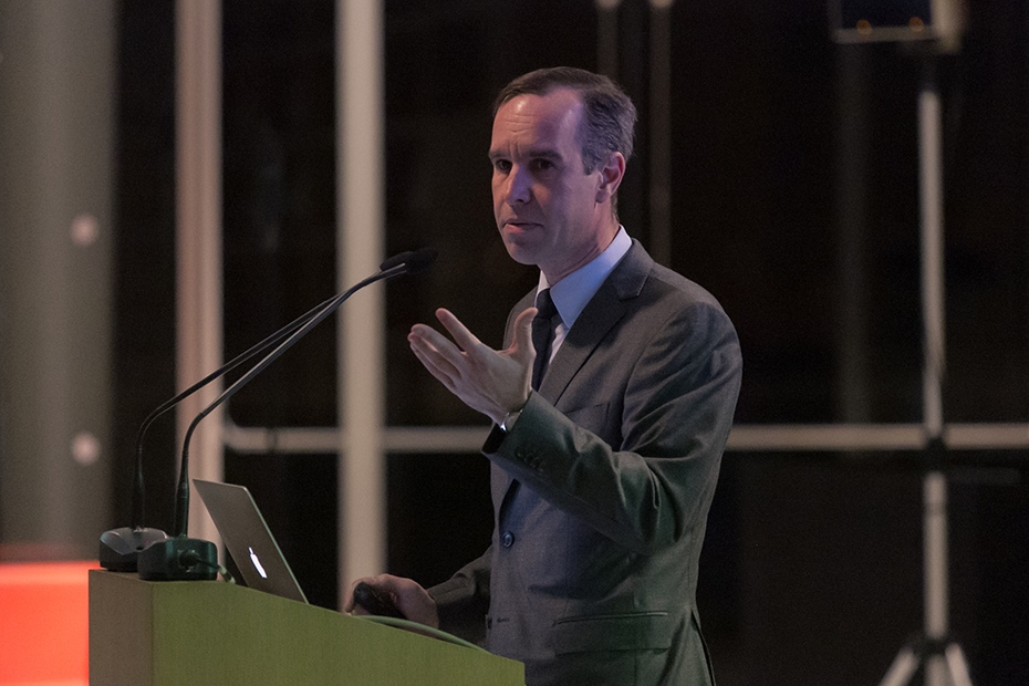 Πλαινή φωτογραφία ενός άντρα που μιλάει στο βήμα. Με το ένα χέρι του χρησιμοποιεί το laptop και με το άλλο εκφράζει τα λόγια του 