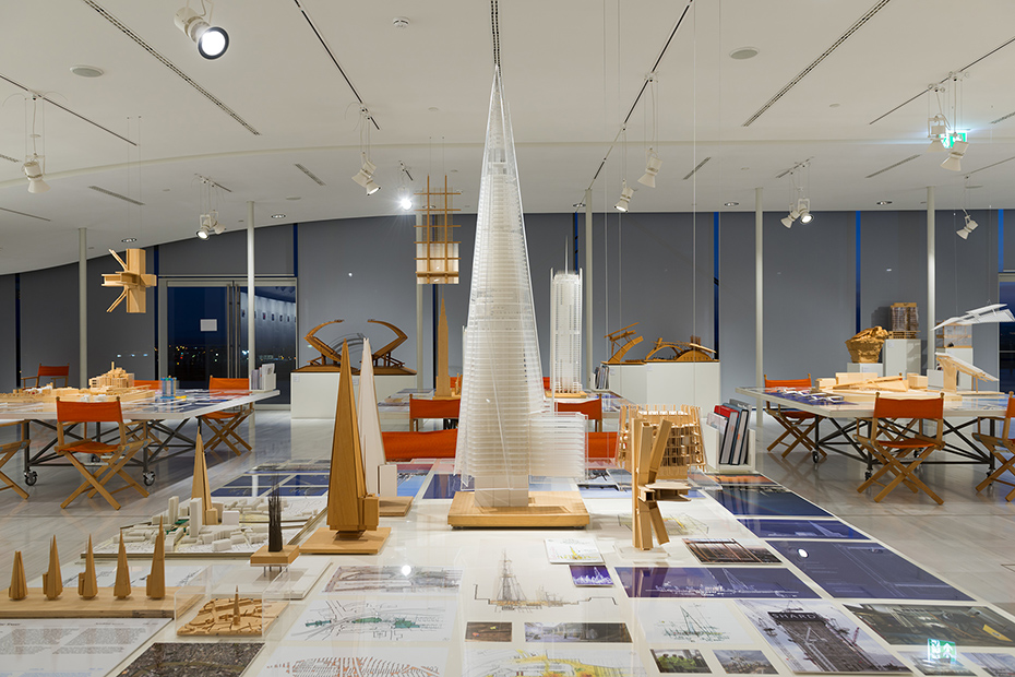 Κοντινή φωτογραφία από ένα τραπέζι στο οποίο υπάρχουν σχέδια, σημειώσεις και μακέτες κτηρίων διαφορετικών μεγεθών και σχεδίων 