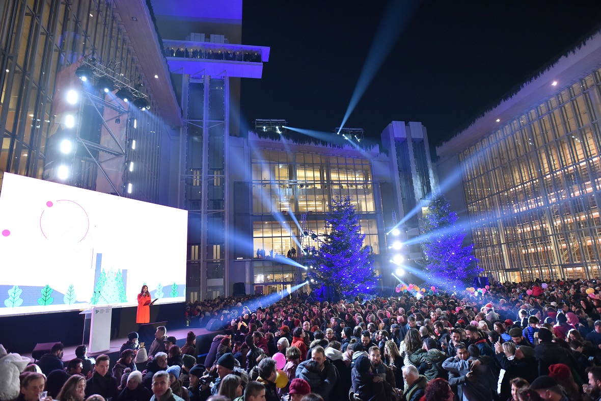 Πλήθος κόσμου στέκεται στον προαύλιο χώρο που περιβάλλεται από παράλληλα κτήρια και παρακολουθούν μια γυναίκα που μιλάει στη σκηνή 