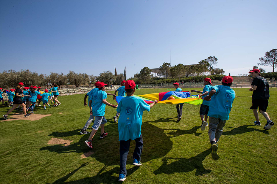 Τα παιδιά κρατούν ένα πολύχρωμο πανί και τρέχουν σε κύκλο σε ένα γήπεδο ποδοσφαίρου.