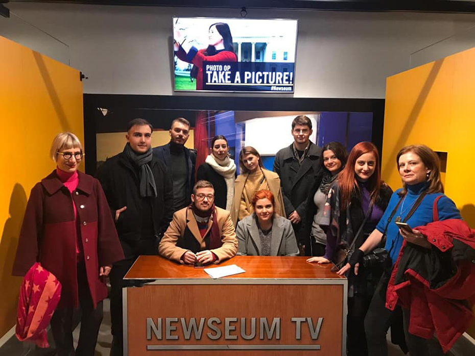Μια ομάδα ανθρώπων στέκεται γύρω από ένα γραφείο που γράφει NEWSEUM TV