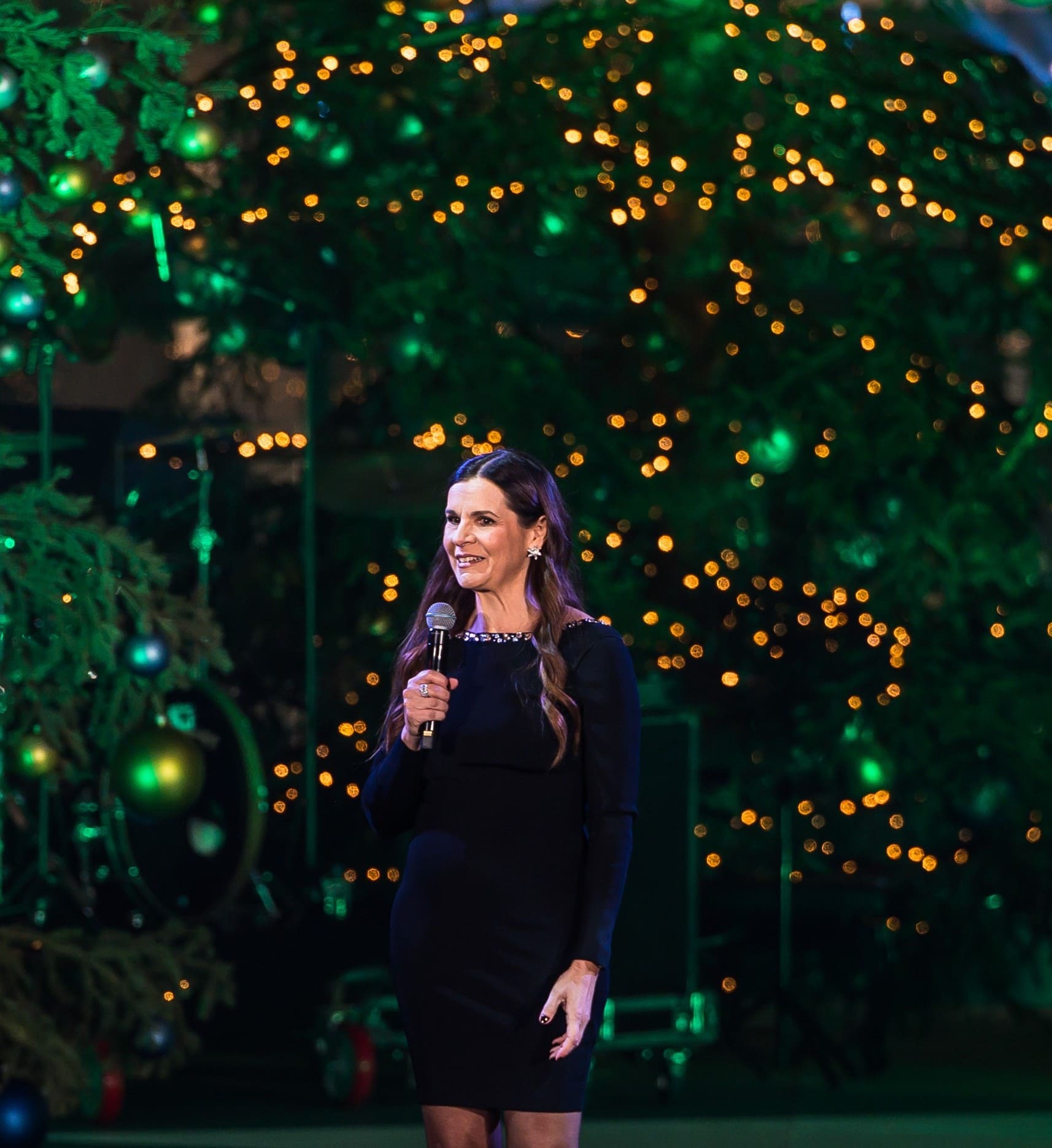 Η παρουσιάστρια στη σκηνή του ΚΠΙΣΝ, μπροστά από χριστουγεννιάτικα δέντρα.