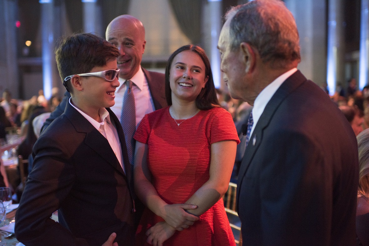 A man talks to two young children behind whom Andreas Dracopoulos, President of the Stavros Niarchos Foundation smiles